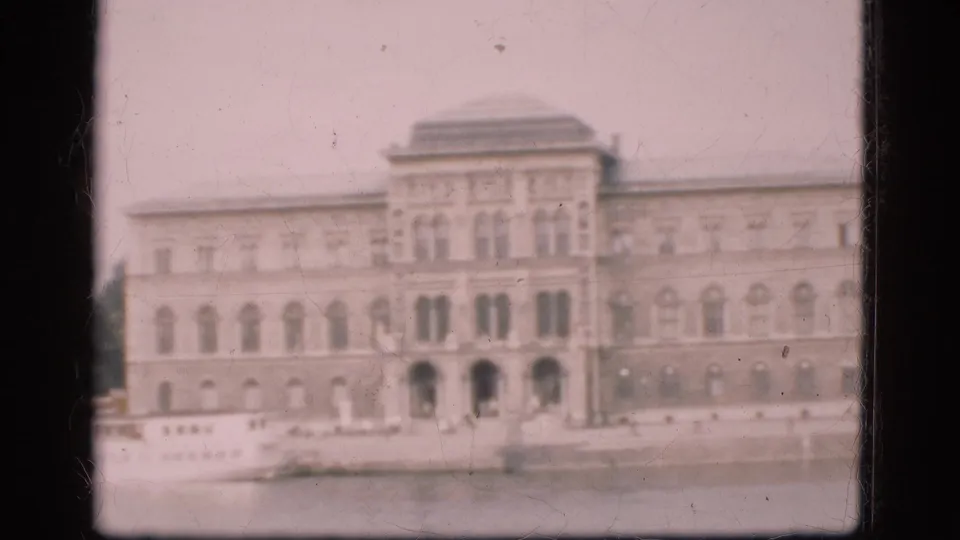 Final 1975 Denmark waterfront view with a broad building facade, arched entrances, small cars, quay, and partial boat visible.