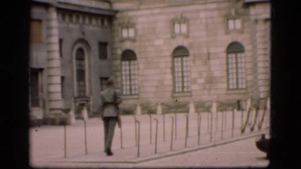 The 1975 Denmark home-movie frame shows the guard from behind, rifle at his side, facing the stone building and arched entrance.