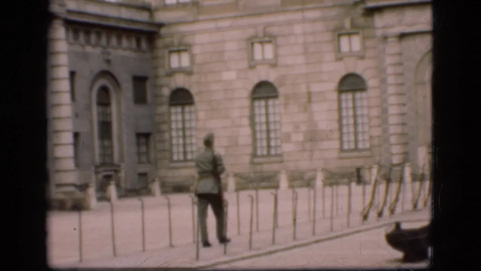 Final 1975 Denmark frame with the uniformed guard standing near bollards before a stone building with tall arched windows.