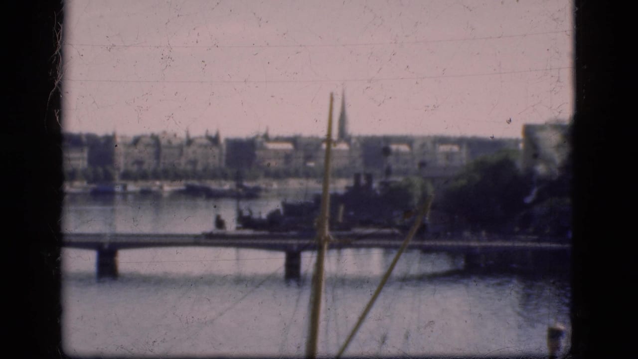 1975 Denmark 8mm frame showing a bridge across calm water, distant shoreline buildings, trees, and a church spire.