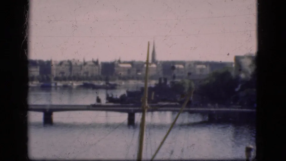 1975 Denmark 8mm frame showing a bridge across calm water, distant shoreline buildings, trees, and a church spire.
