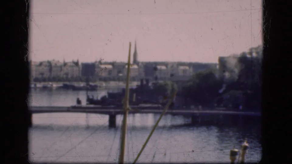 1975 Denmark 8mm frame with a bridge over water, distant buildings, trees, and a church spire behind dark film edges.