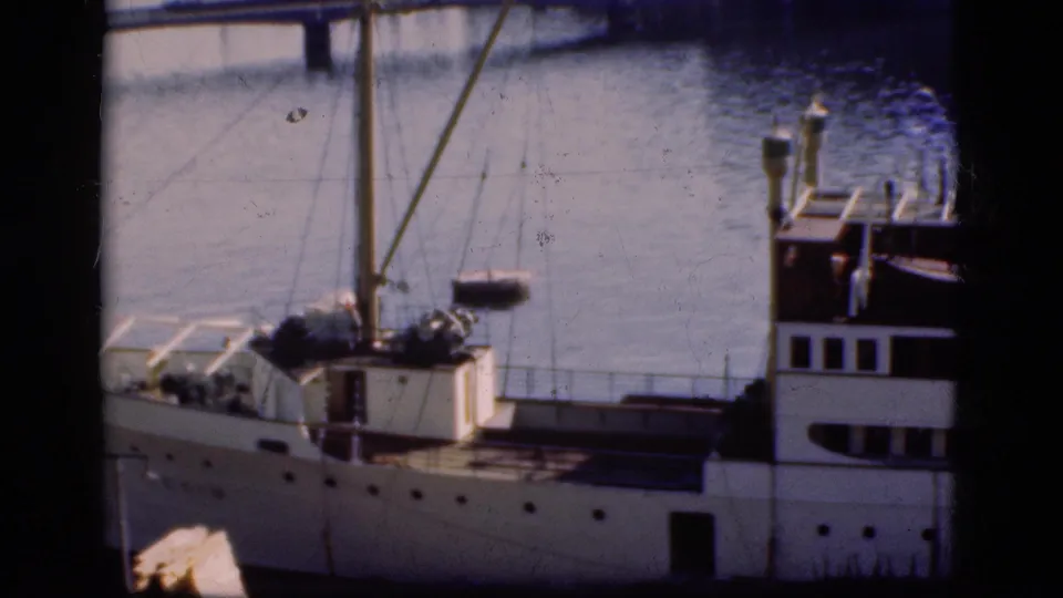 1975 Denmark home-movie frame showing a white docked passenger ship, mast rigging, railings, and calm harbor water.