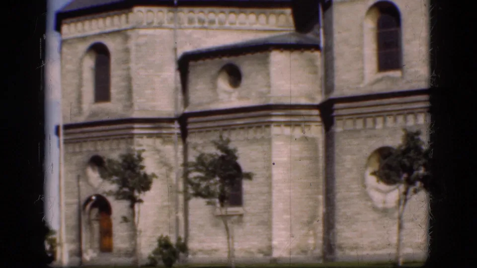 1975 Denmark home-movie frame showing a historic stone building facade with arched windows, round openings, and trees.