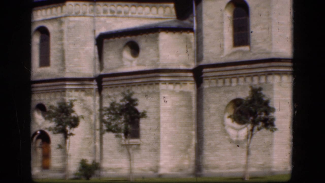 Historic pale stone facade with arched windows, small trees, grass, and dark film borders.