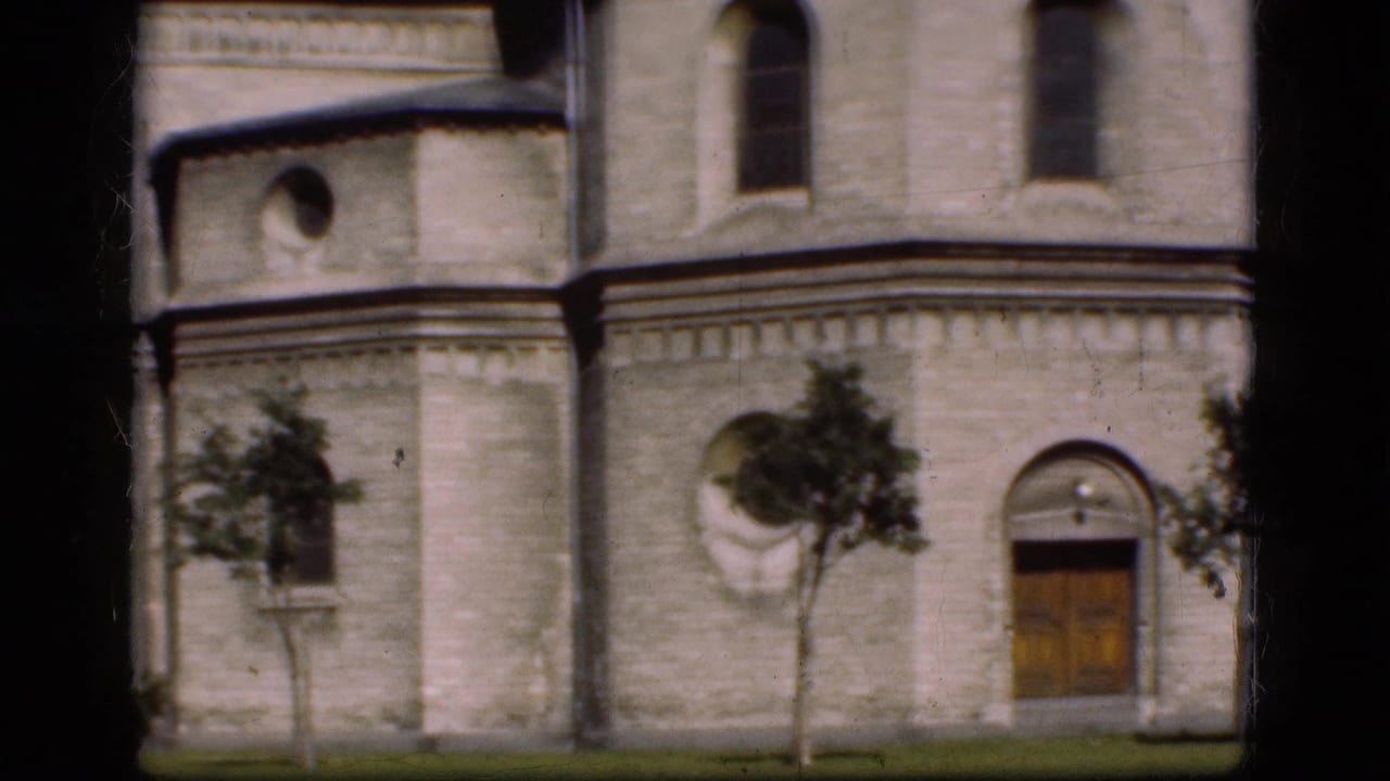 Stone building facade with arched doorway, wooden door, arched windows, trees, and lawn.