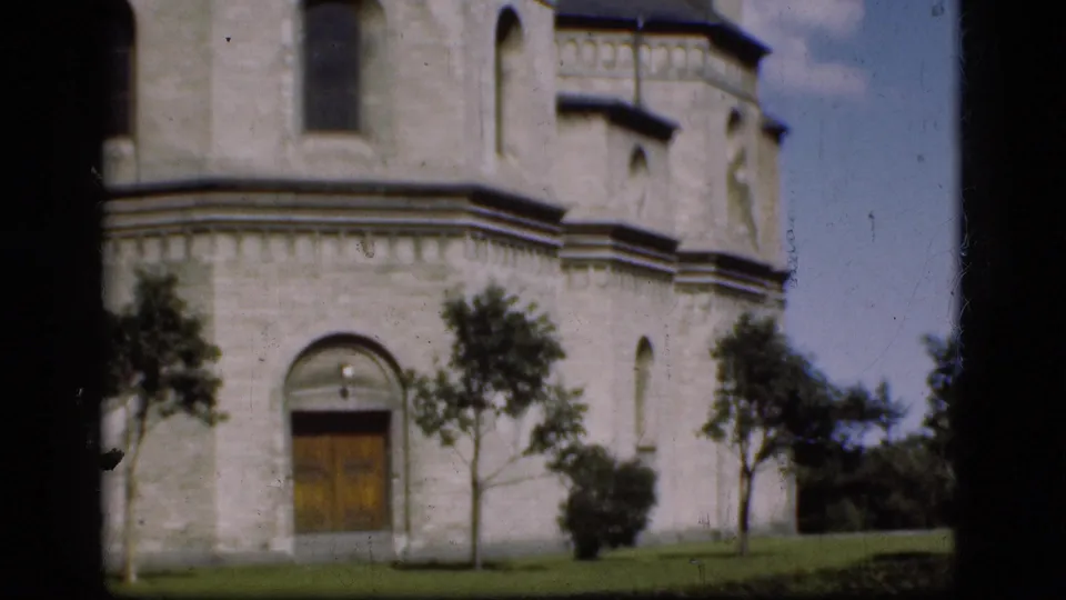 1975 Denmark 8mm frame showing the pan reaching a building corner with doorway, trees, lawn, and sky.