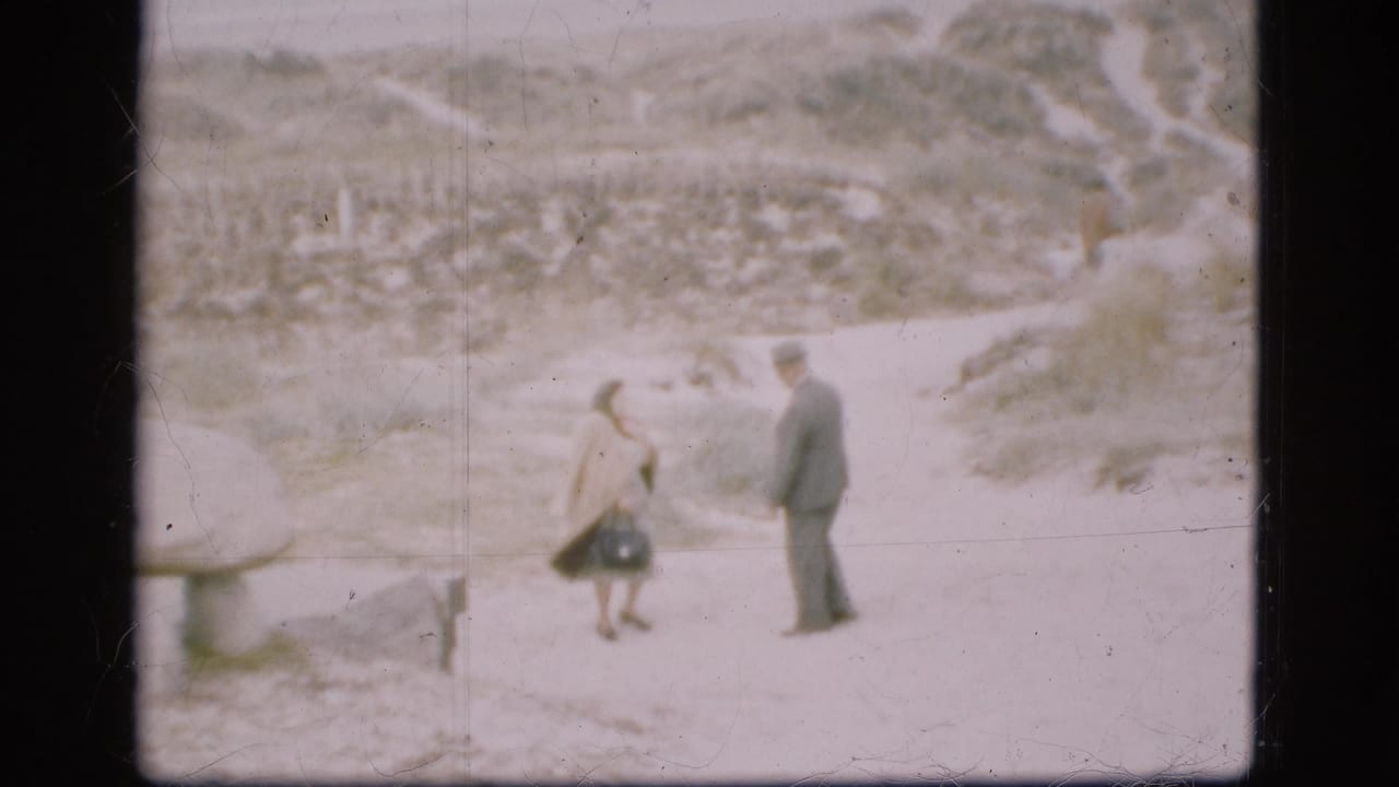 A man in a dark suit and hat faces a woman in a light coat on a sandy path at an outdoor Danish historic site.