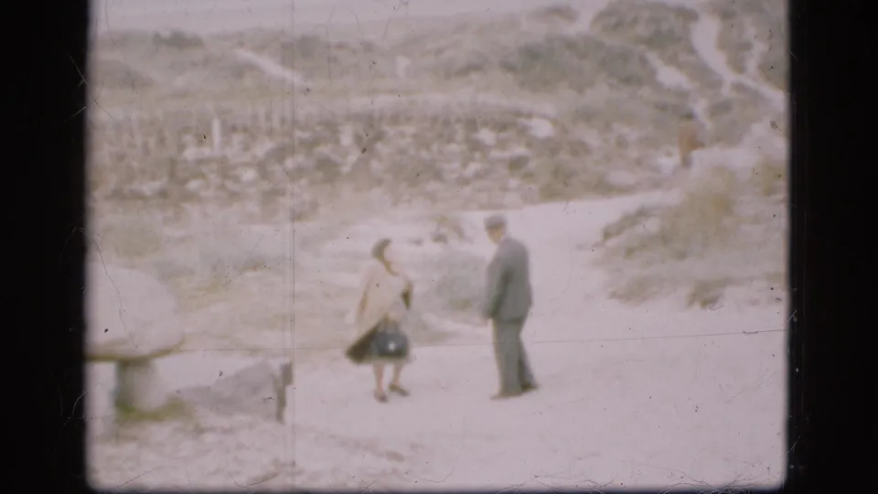 A man in a dark suit and hat faces a woman in a light coat on a sandy path at an outdoor Danish historic site.
