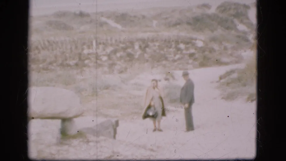 The couple remain near a low stone form, with the woman carrying a handbag and the man in dark formal clothing.