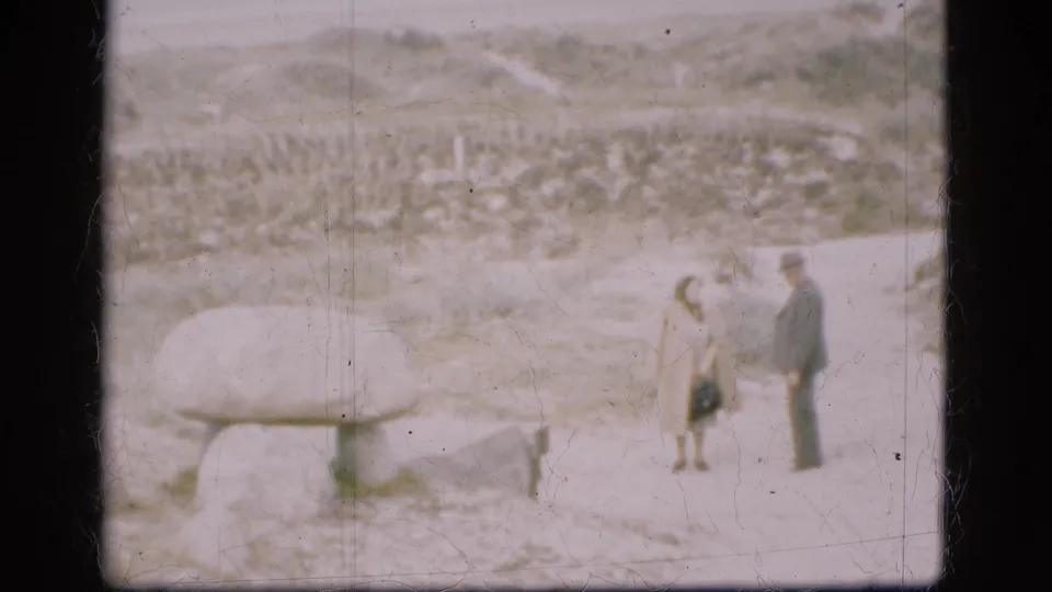 A 1975 Denmark home-movie frame shows the couple beside a broad low stone structure, with rough terrain and pale background forms behind them.