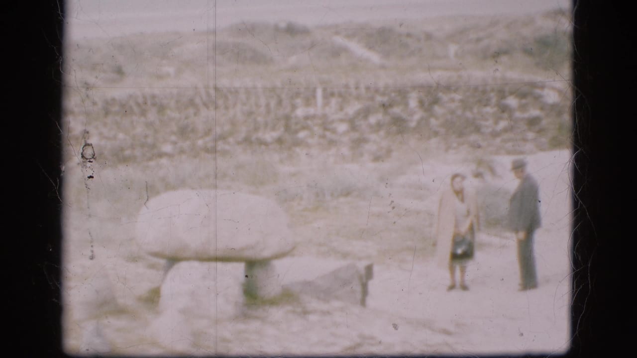In Denmark 1975 home-movie footage, two adults pause beside a large stone monument on snow-covered ground.