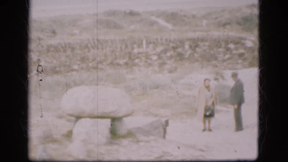 In Denmark 1975 home-movie footage, two adults pause beside a large stone monument on snow-covered ground.