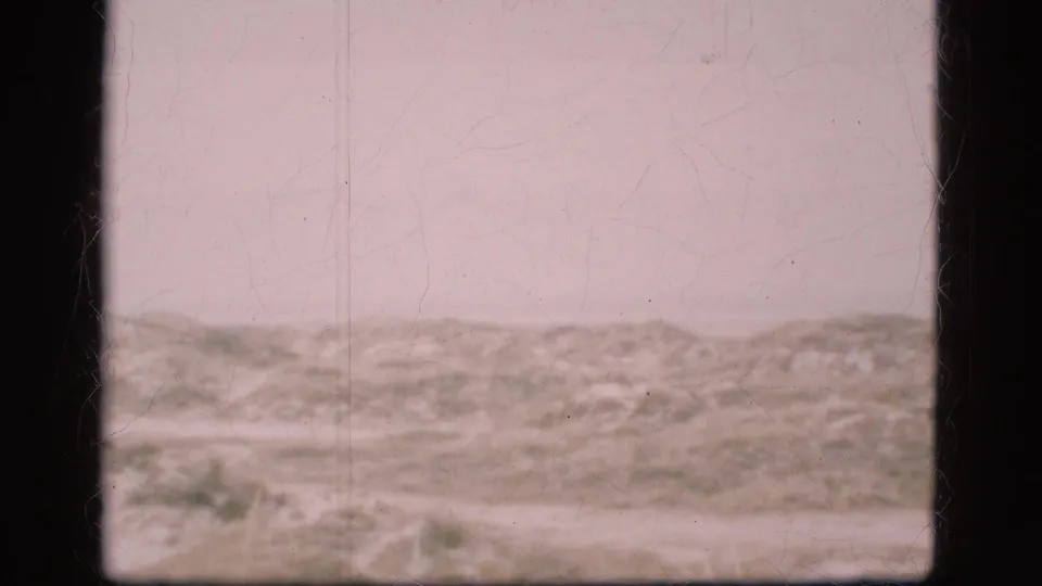 1975 Danish home-movie frame of sparse open terrain and heavy cloud, with fading, scratches, and a soft 8mm scan texture.