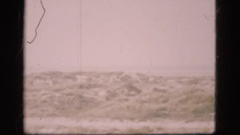 1975 Denmark 8mm frame with sandy scrub terrain, a faint horizontal track, pale sky, and visible age marks from the film scan.