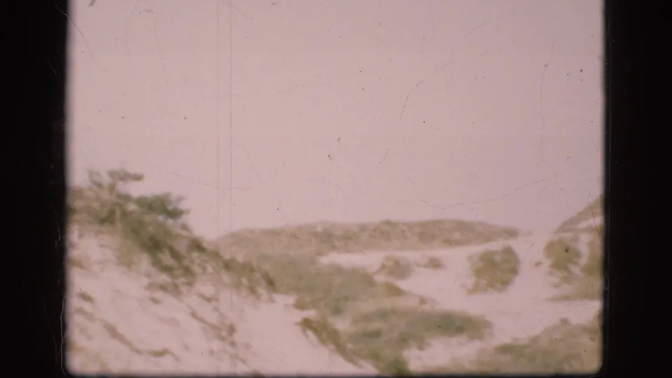 1975 Danish home-movie frame of snow-covered dunes or low hills, with scrub vegetation at the edges and visible 8mm wear.