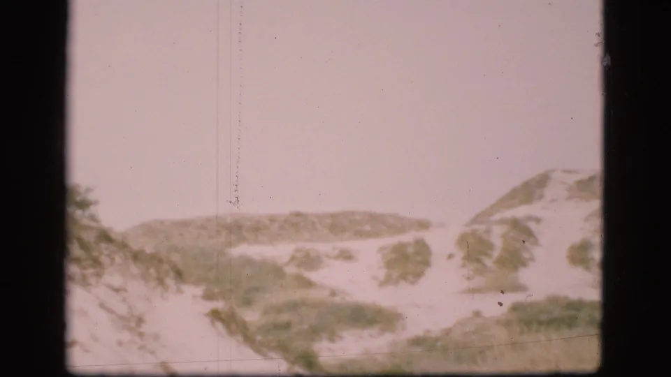 Grainy 8mm frame from Denmark in 1975 showing a barren snowy landscape with low hills, sparse greenery, and film gate borders.