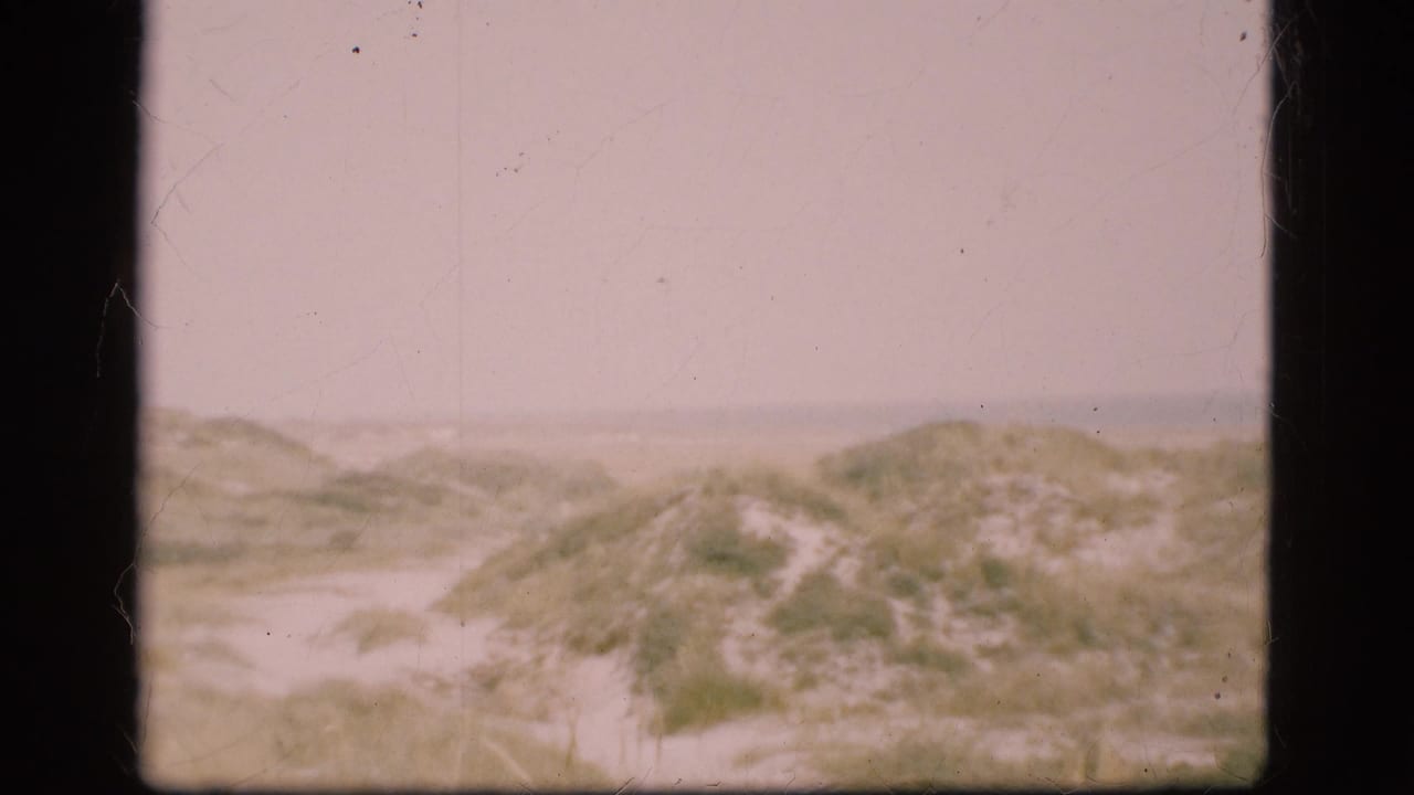 1975 Denmark home-movie frame showing low sandy dunes, sparse vegetation, and a faint path beneath an overcast sky.