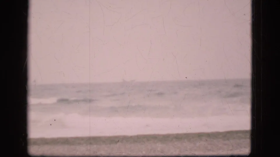 1975 Denmark 8mm frame showing grey-blue surf and a dark beach strip under a pale overcast sky.