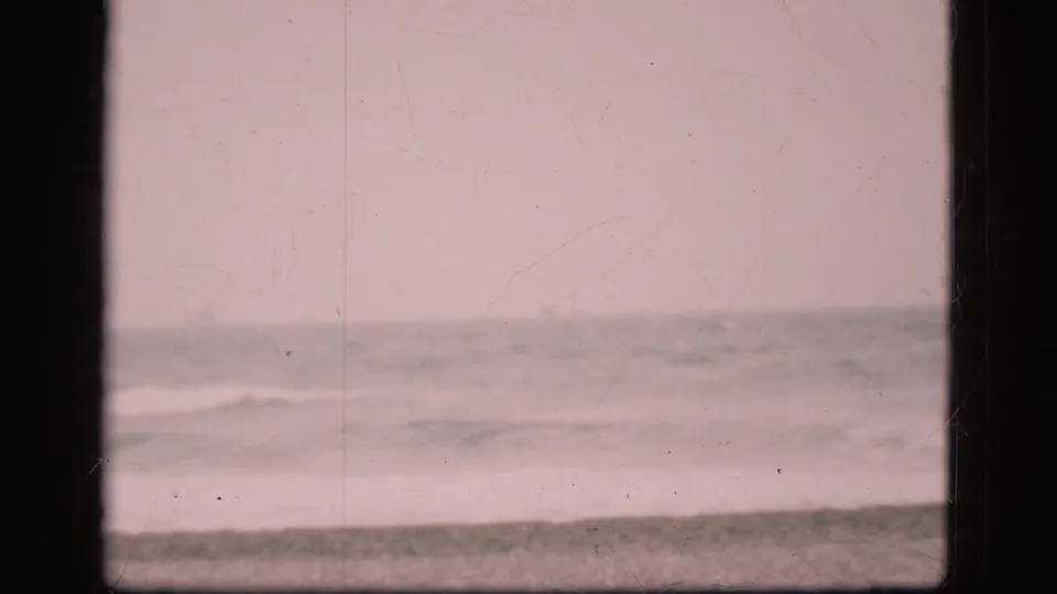 Soft 1975 Denmark shoreline frame with low waves, muted sea color, and beach foreground in visible 8mm texture.