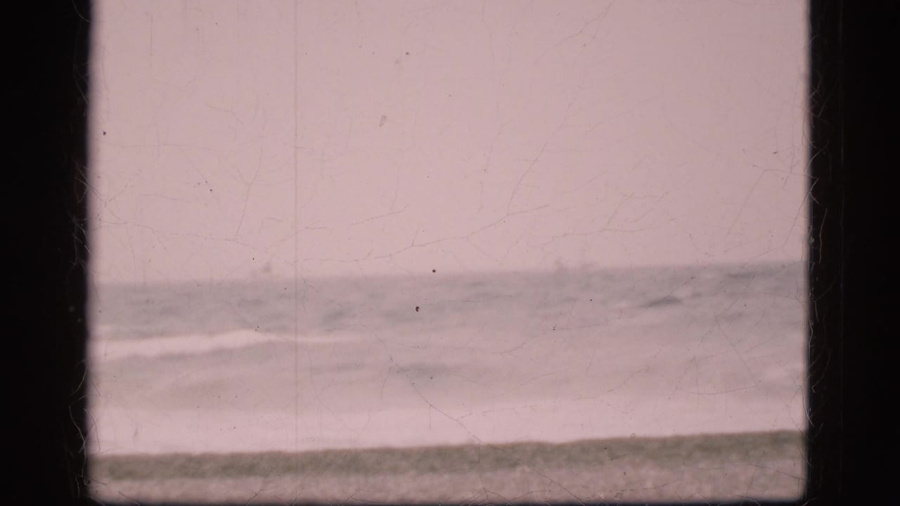 1975 Danish beach view with pale cloudy sky, distant ocean horizon, gentle surf, and visible 8mm film borders and wear.
