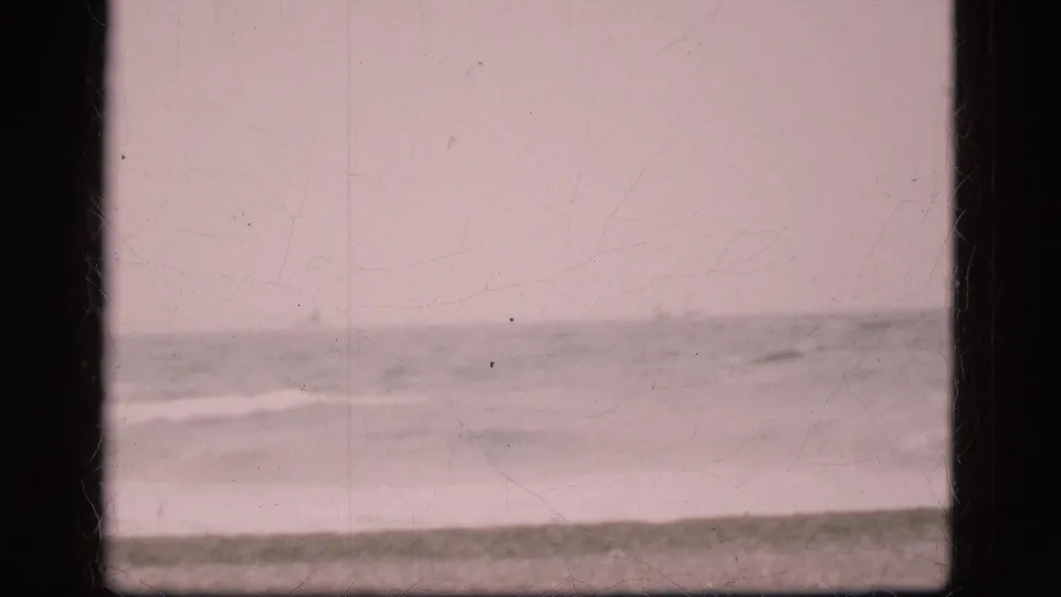 1975 Danish beach view with pale cloudy sky, distant ocean horizon, gentle surf, and visible 8mm film borders and wear.