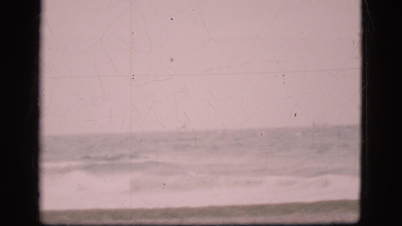 Empty Danish coastal view from 1975 showing low waves, cloudy sky, muted color, and visible scratches on the scanned 8mm frame.