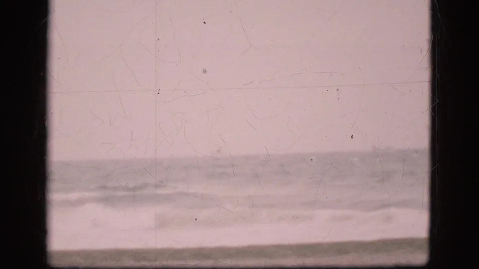 Empty Danish coastal view from 1975 showing low waves, cloudy sky, muted color, and visible scratches on the scanned 8mm frame.