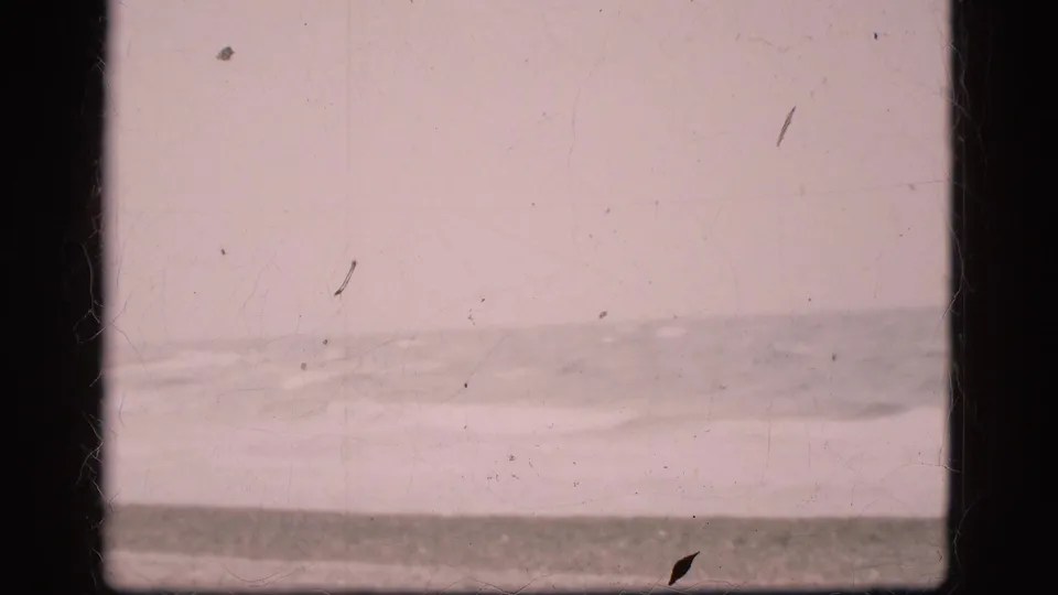 1975 Denmark beach footage showing an empty shoreline, soft surf, pale overcast sky, dark film borders, and visible dust marks.