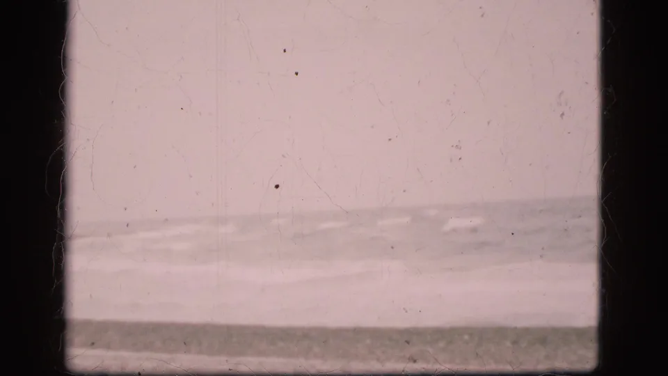 Danish 1975 8mm frame of a quiet empty beach, low wave line, cloudy sky, soft exposure, and aged-film surface marks.