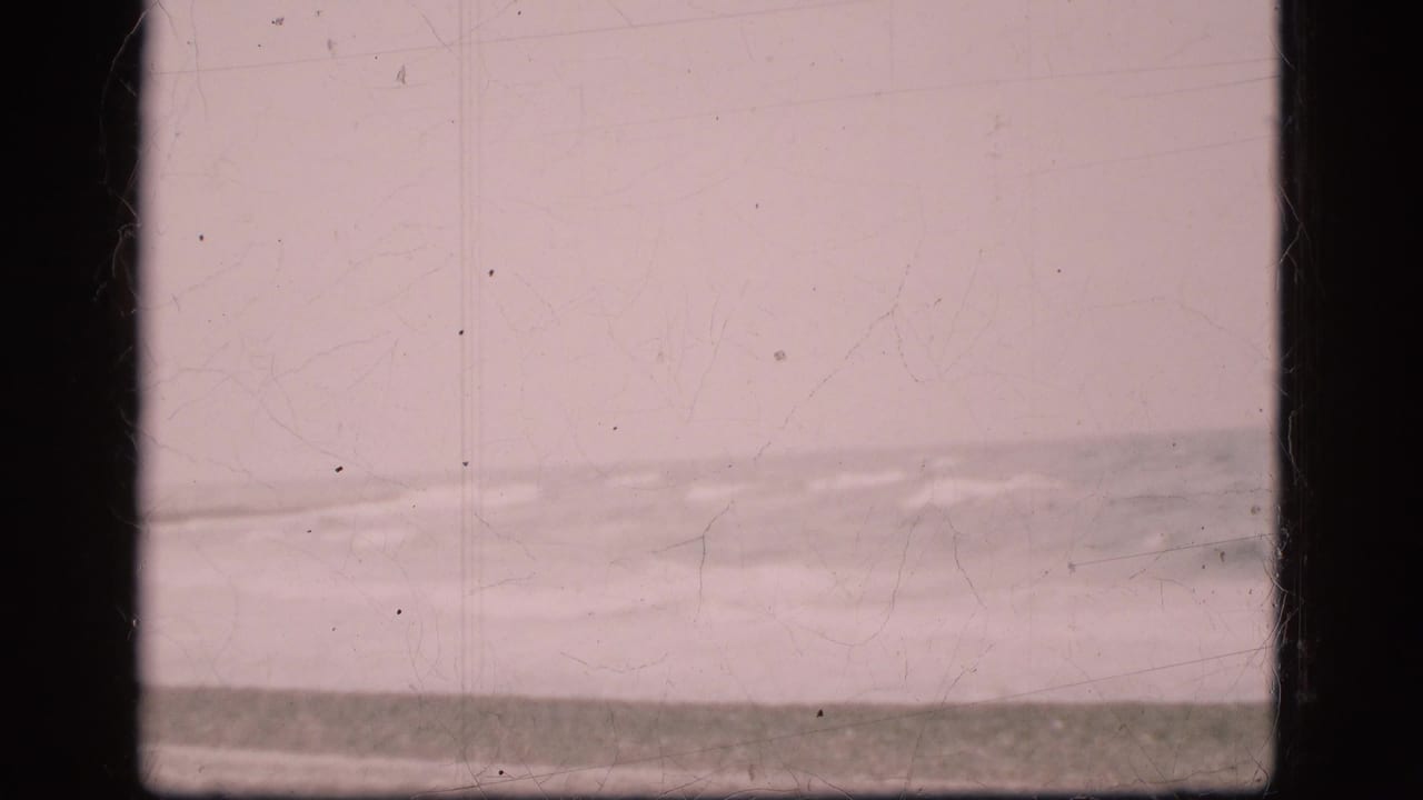 1975 Danish coastal home-movie frame with low waves breaking near a sandy beach beneath an overcast sky.