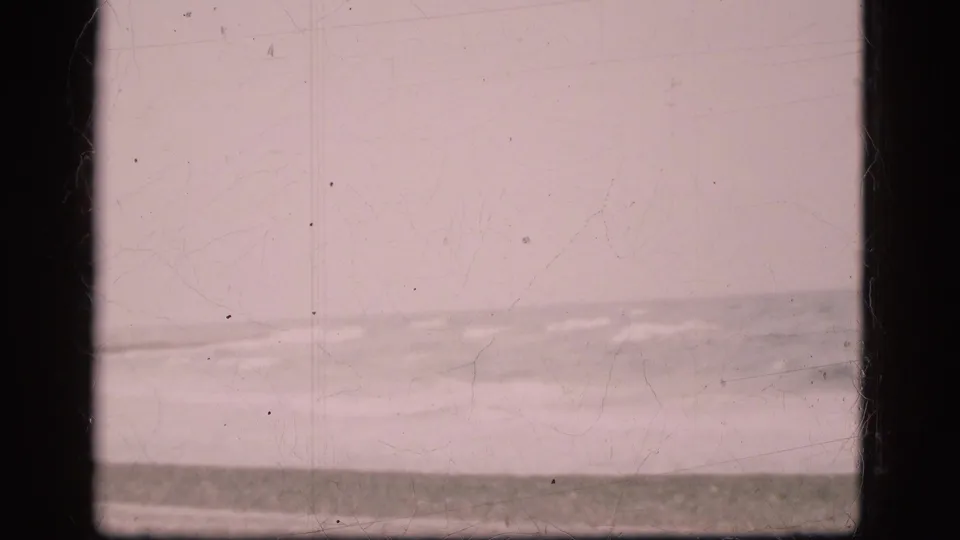 1975 Danish coastal home-movie frame with low waves breaking near a sandy beach beneath an overcast sky.