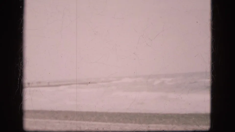 Faded 1975 home-movie frame of waves and beach in Denmark, with visible grain and light surface wear.