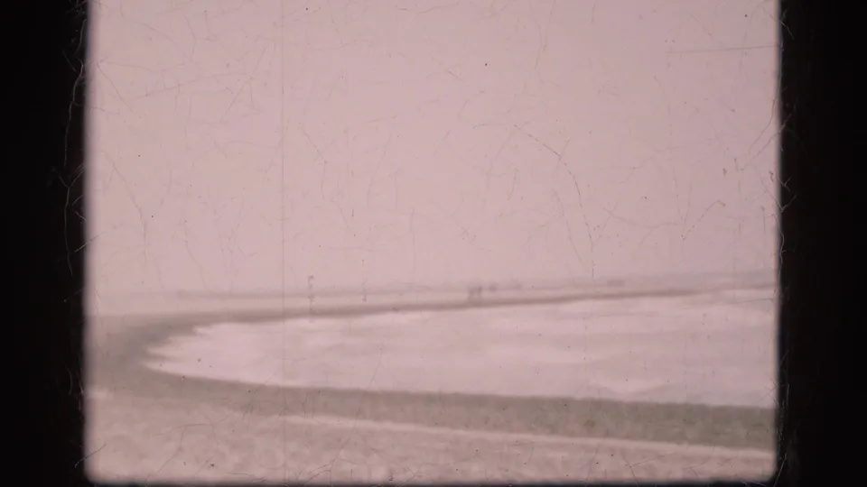 1975 Denmark 8mm frame with a curving surf edge, low beach shoreline, pale sky, and visible dust and scratches from the film scan.