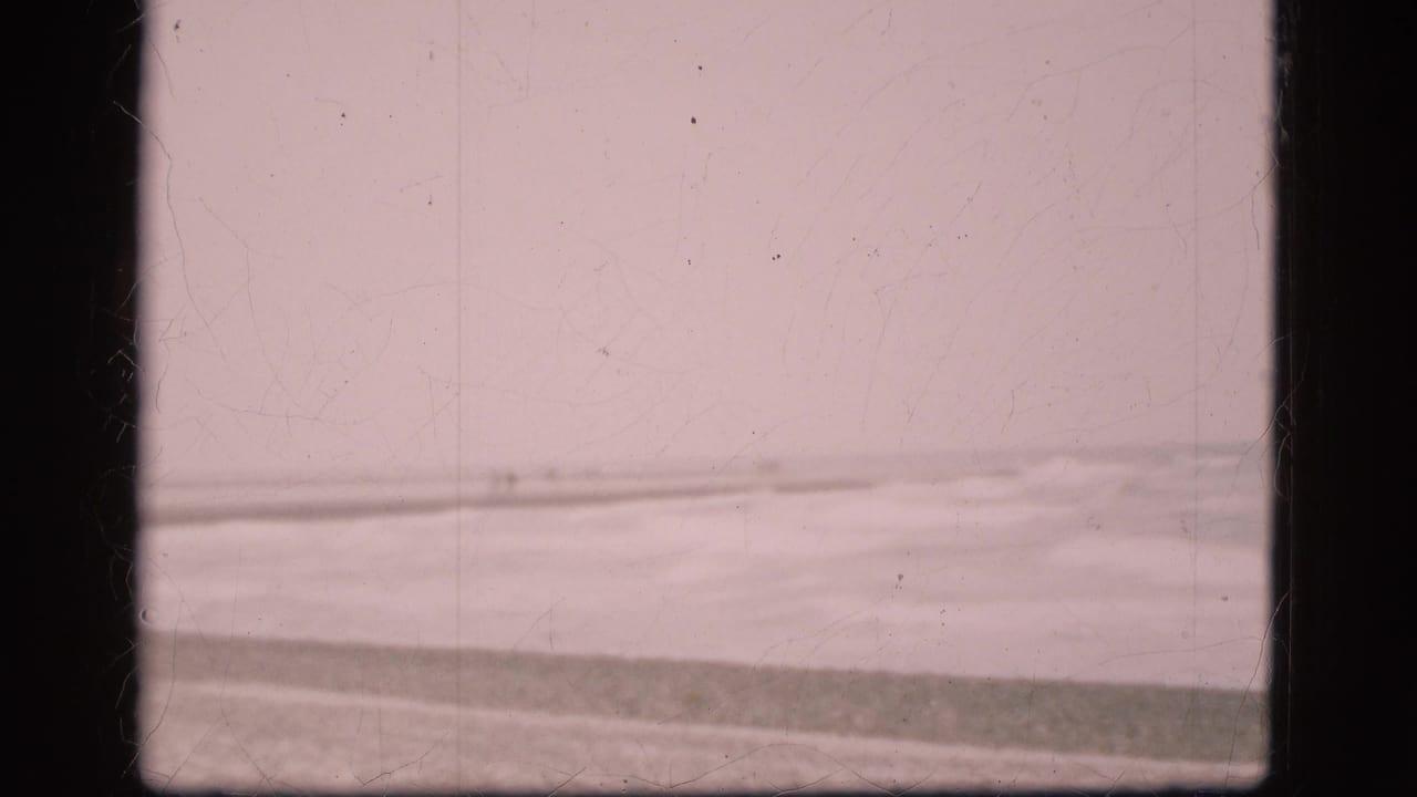 Faded 8mm frame of a Danish beach in 1975, showing pale surf, pebbles, and an overcast horizon through film scratches.