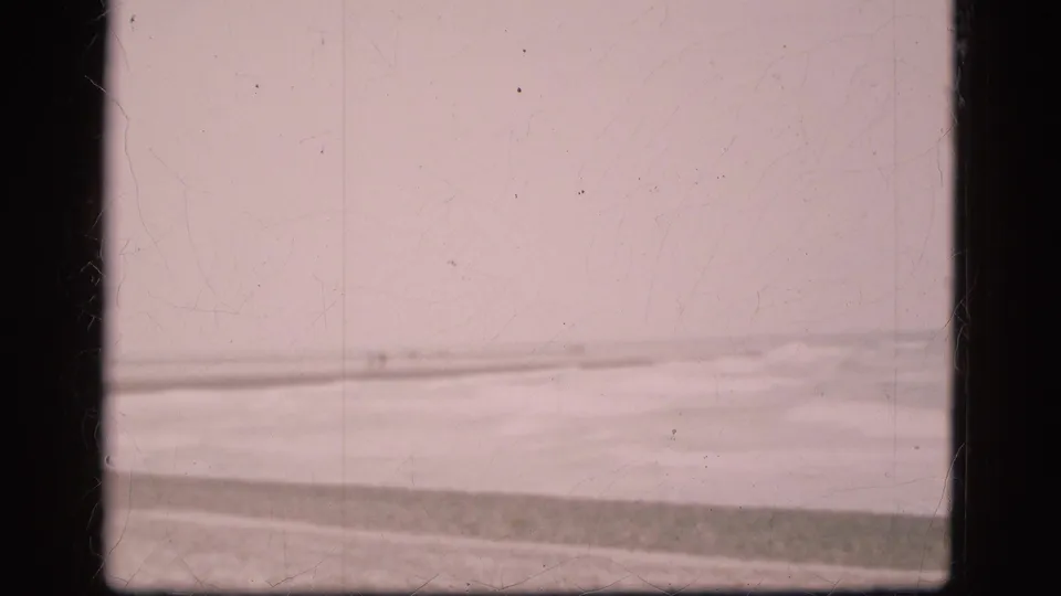 Faded 8mm frame of a Danish beach in 1975, showing pale surf, pebbles, and an overcast horizon through film scratches.