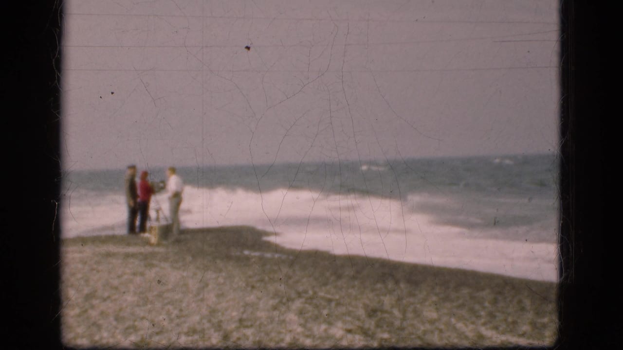 Three distant figures stand together on a pebbled Danish shore in 1975, facing the surf in a grainy 8mm home-movie frame.