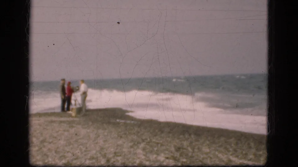 Three distant figures stand together on a pebbled Danish shore in 1975, facing the surf in a grainy 8mm home-movie frame.