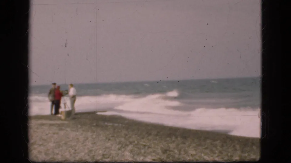 Grainy 1975 Denmark beach frame with three distant people near the surf line, pebbles in the foreground, and muted sea beyond.
