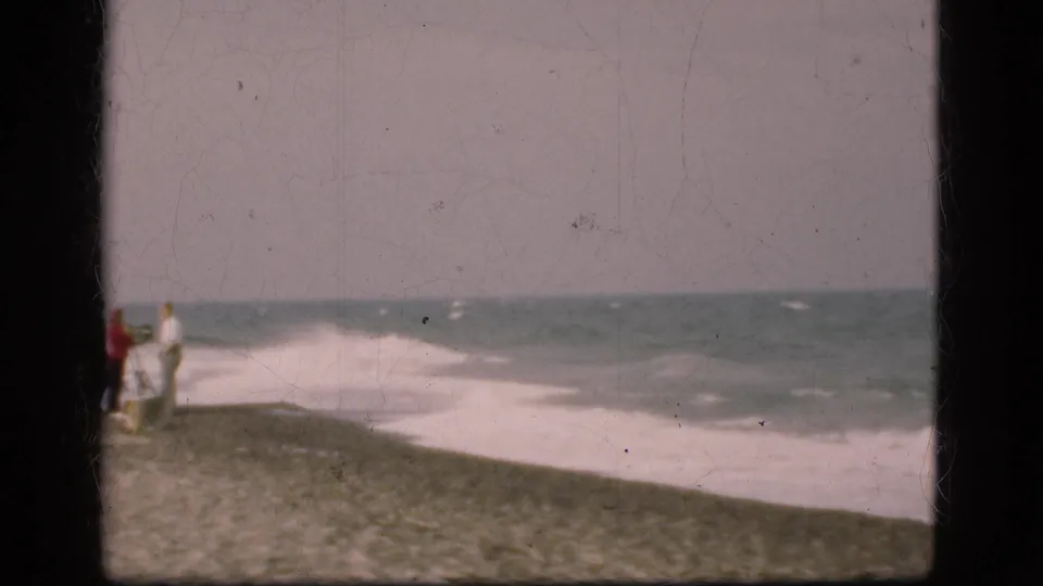 Cropped 8mm frame from a 1975 Danish beach visit, showing the group at frame edge and rough surf across the pebbled shore.