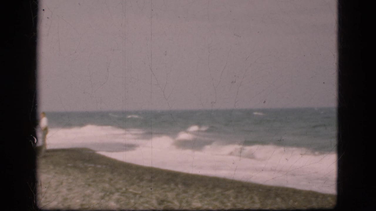 1975 Denmark 8mm frame showing surf breaking along a sloped sandy shoreline under a gray sky.