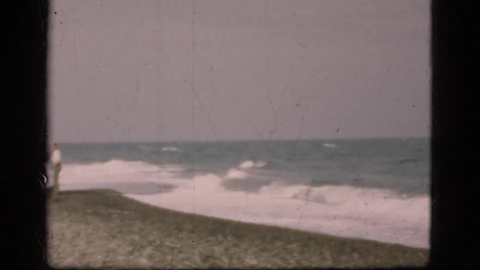 1975 Denmark 8mm frame showing surf breaking along a sloped sandy shoreline under a gray sky.