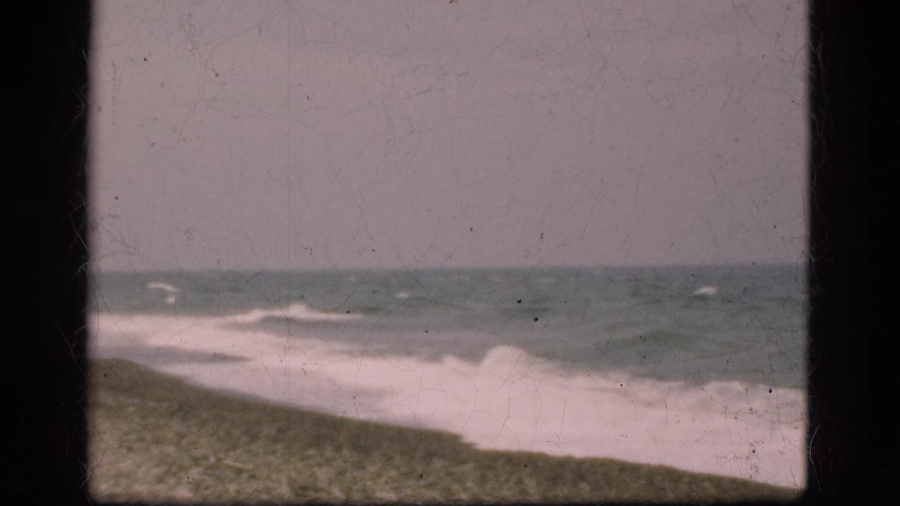 1975 Denmark beach view with muted blue-gray water, low surf, sandy foreground, and visible 8mm frame edges.