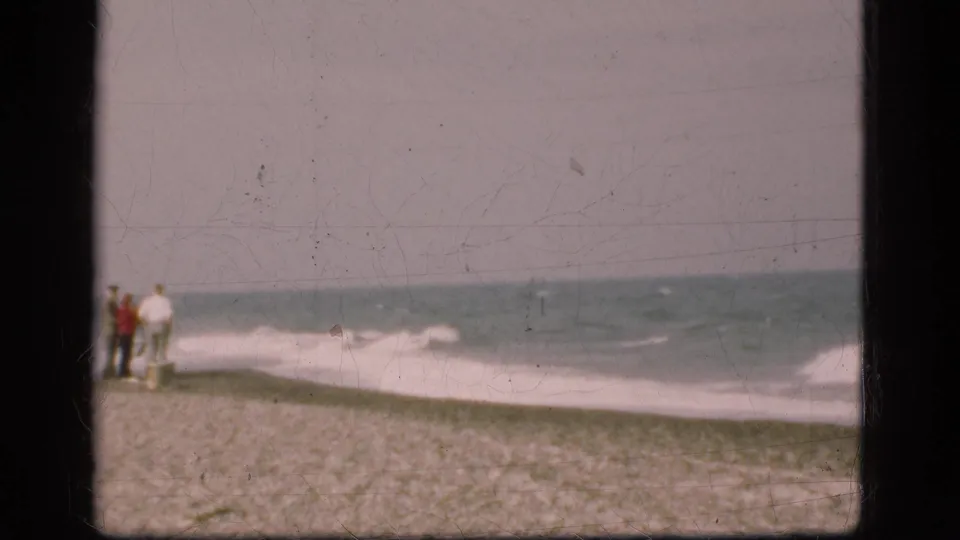 1975 Denmark 8mm frame with distant people standing on sand and looking toward waves along the shoreline.