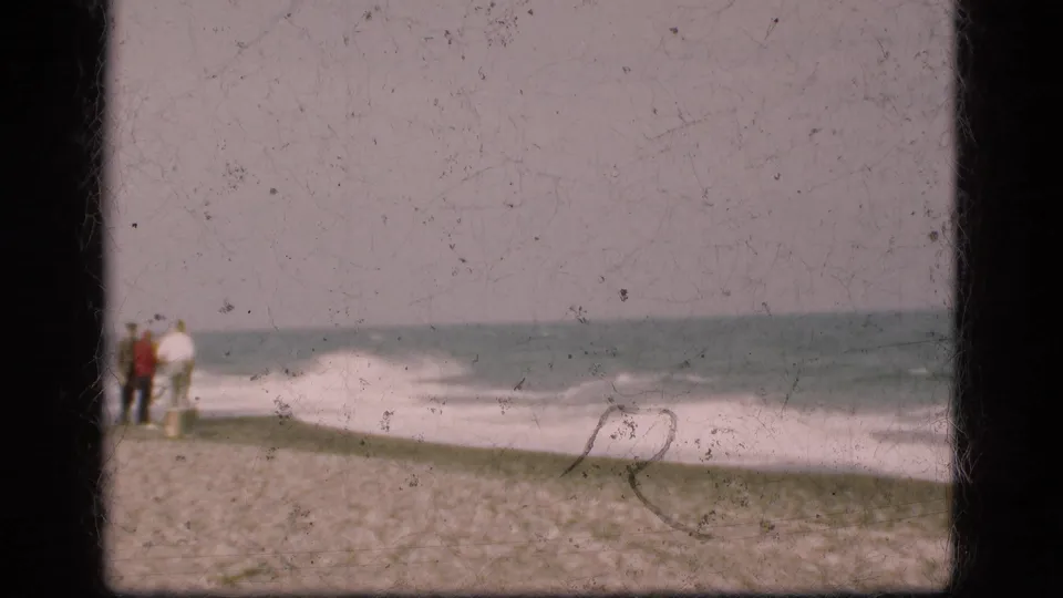 1975 Denmark coastal home-movie frame showing distant figures on sand beside rolling surf and a muted horizon.