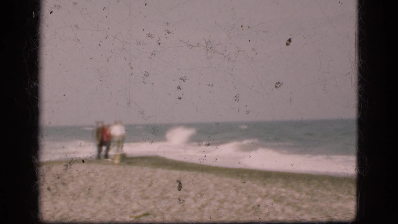 Distant figures stand on a Danish coastal strip as surf rolls in, seen in soft 1970s 8mm home-movie footage.