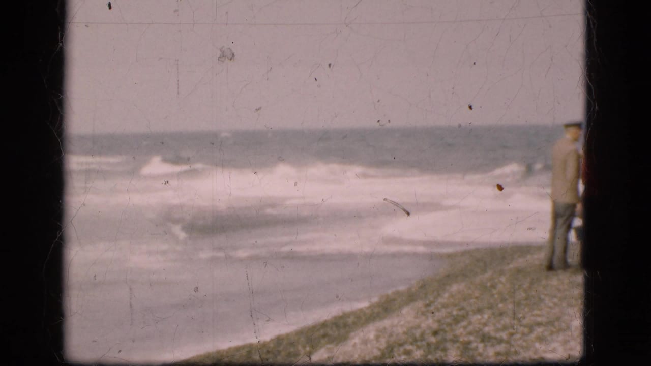 Waves roll against a Danish beach while one distant figure is partly framed at the edge in faded 8mm footage.