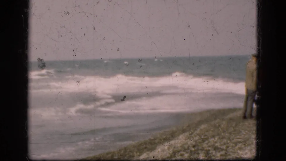 A lone distant figure stands near the Danish shoreline, watching surf in a soft, scratched 1970s 8mm frame.