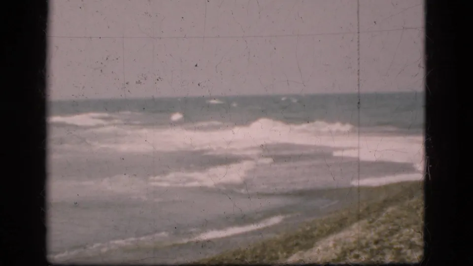 The Danish coast appears without clear figures, showing churning surf and a sloped beach in faded 1970s 8mm color.
