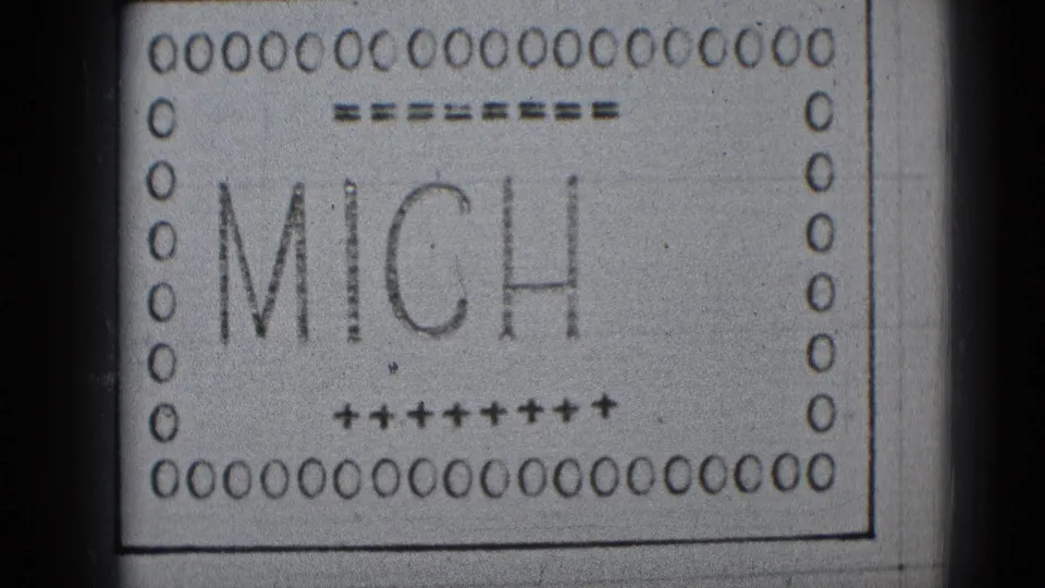 A monochrome 8mm leader frame shows the partial name MICH, surrounded by perforations, plus signs, dashes, and film wear.